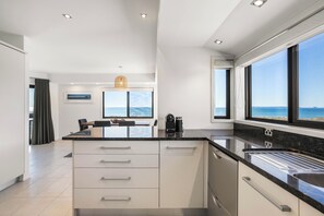 A modern kitchen with a large island, a dining area with a view, and a kitchen island with a built-in dishwasher. - Sunbrae Sands - Mount Maunganui Holiday Homes (Mount Maunganui)