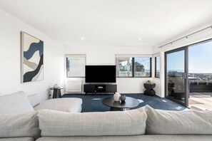 A modern living room with a flat-screen TV, a sofa, a coffee table, and a large window offering a view of the city. - Sunbrae Sands - Mount Maunganui Holiday Homes (Mount Maunganui)