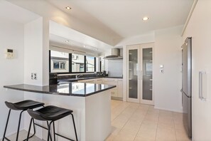 Eine moderne Küche mit einer zentralen Kochinsel, Barhocker, einem Edelstahlkühlschrank und Blick ins Freie durch große Fenster.