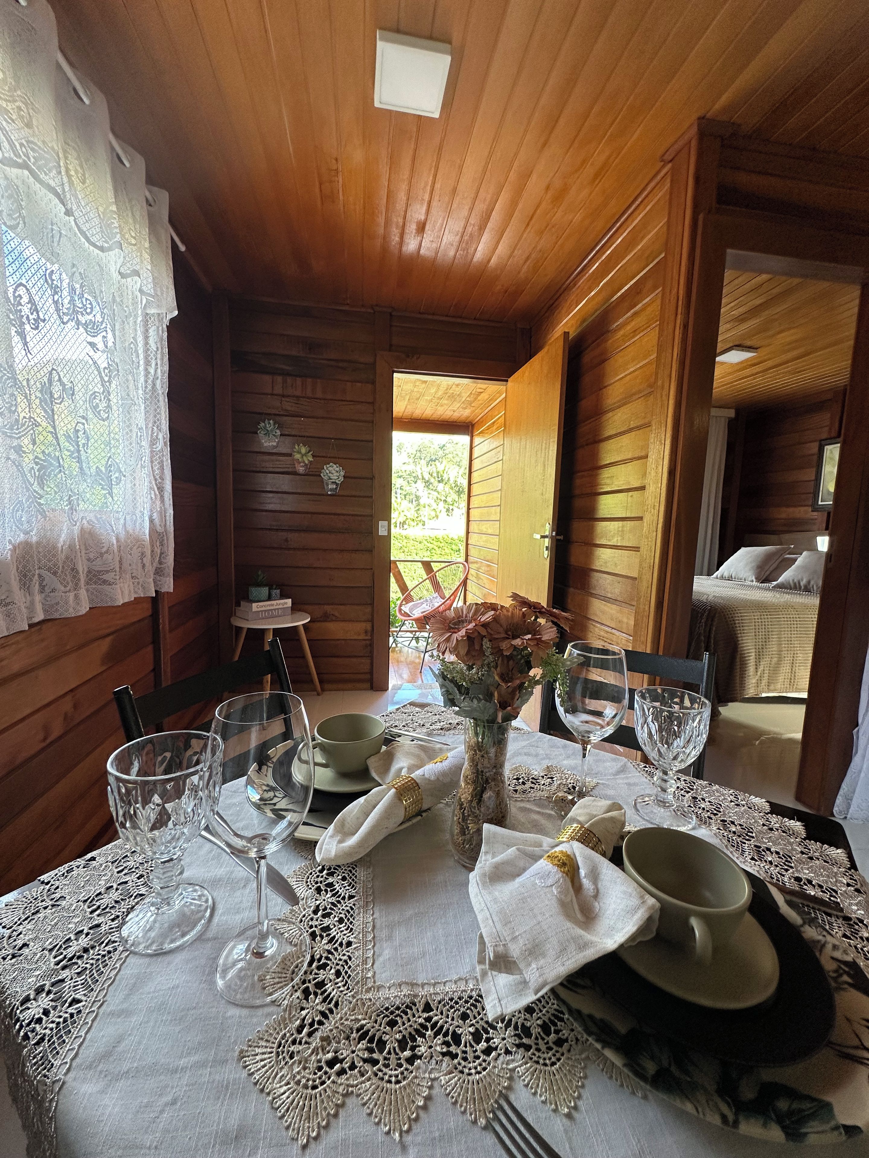 Interior de una cabaña de madera con una mesa para dos, mantel individual de encaje, un florero con flores y vistas al exterior.