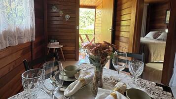 Interior de una cabaña de madera con una mesa para dos, mantel individual de encaje, un florero con flores y vistas al exterior.