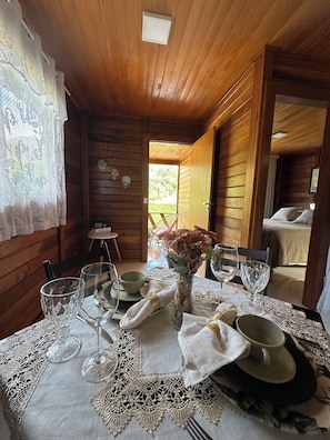 Interior de una cabaña de madera con una mesa para dos, mantel individual de encaje, un florero con flores y vistas al exterior.