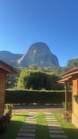 Vista de una montaña con un globo aerostático a lo lejos, un jardín cuidado con un camino, y una estructura de madera con un porche.