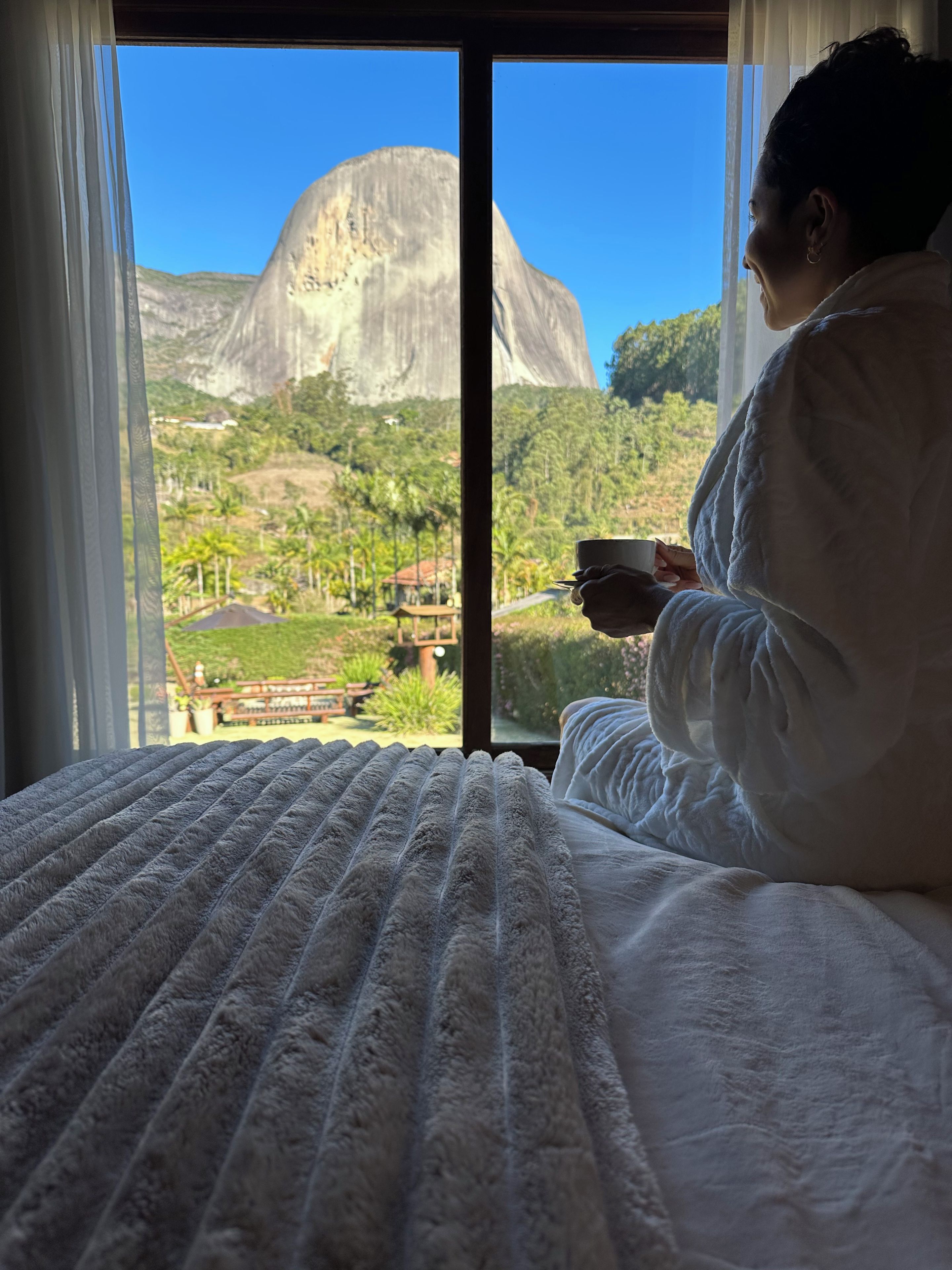 Una persona sentada en una cama con vista a una gran montaña y vegetación.