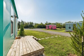 A wooden deck leading to a building with a green exterior, a red structure in the background, and a clear sky. - Modern Boxabl Casita | Stillwater OK | Pasadera 5 (Stillwater)