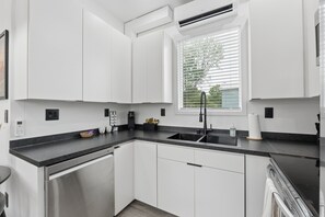 A modern kitchen with white cabinets, a black countertop, and a window with blinds. - Modern Boxabl Casita | Stillwater OK | Pasadera 5 (Stillwater)