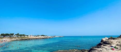 A coastal beach scene with clear blue water, rocky shoreline, and distant buildings.
