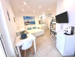 Quarto de hotel compacto com cama, escrivaninha com cadeira, mesinha com planta, TV fixada na parede e mini-geladeira.