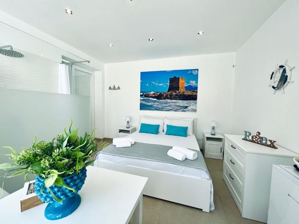 Quarto de hotel com cama, escrivaninha, cadeira, chuveiro e uma pintura com paisagem costeira.