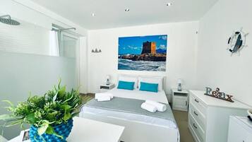 Quarto de hotel com cama, escrivaninha, cadeira, chuveiro e uma pintura com paisagem costeira.