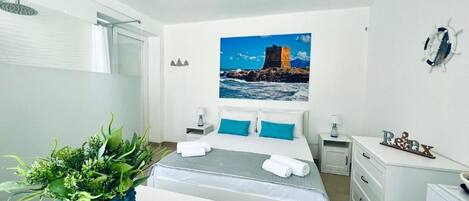 A hotel room with a bed, a desk, a chair, a shower, and a painting of a coastal scene.