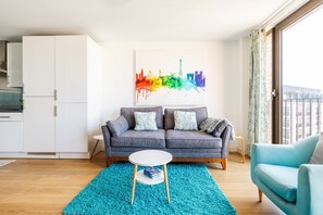 Un salon moderne avec un canapé gris, une petite table blanche et un fauteuil bleu. Une œuvre d’art représentant un paysage urbain coloré est accrochée au mur.