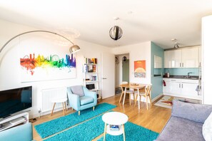 Un salon moderne avec un téléviseur à écran plat, un fauteuil bleu, une table à manger avec des chaises, une bibliothèque et un tableau représentant un paysage urbain aux couleurs vives.