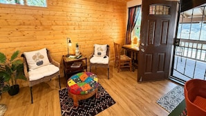 A cozy room with wooden walls, a round ottoman, two chairs, a small table with a lamp, and a door leading outside. - Adorable, easy access, level cabin super clean! (Ruidoso)