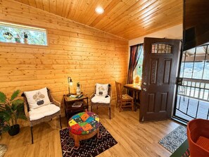 Ein gemütlicher Raum mit Holzwänden, einem runden Ottomanen, zwei Stühlen, einem kleinen Tisch mit Lampe und einer Tür, die nach draußen führt.