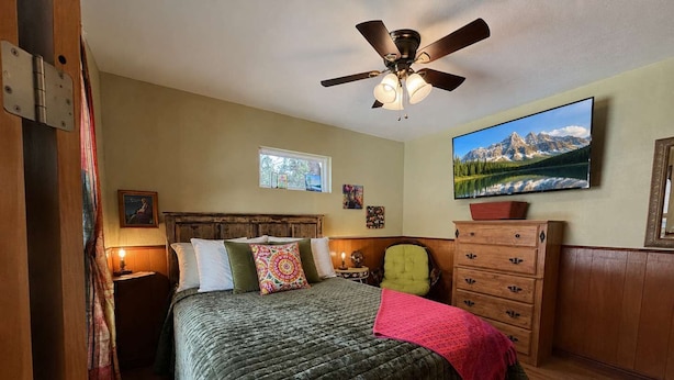 A bedroom with a bed, a ceiling fan, a dresser, a television, and artwork on the wall. - Adorable, easy access, level cabin super clean! (Ruidoso)