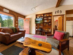 A cozy living room with a brown leather couch, a wooden coffee table, and a grey armchair. A bookshelf filled with various items is against the wall, and a pink door is visible. - Canyon Cabin w/ Hot Tub, Mountain Views & Privacy (Ruidoso)