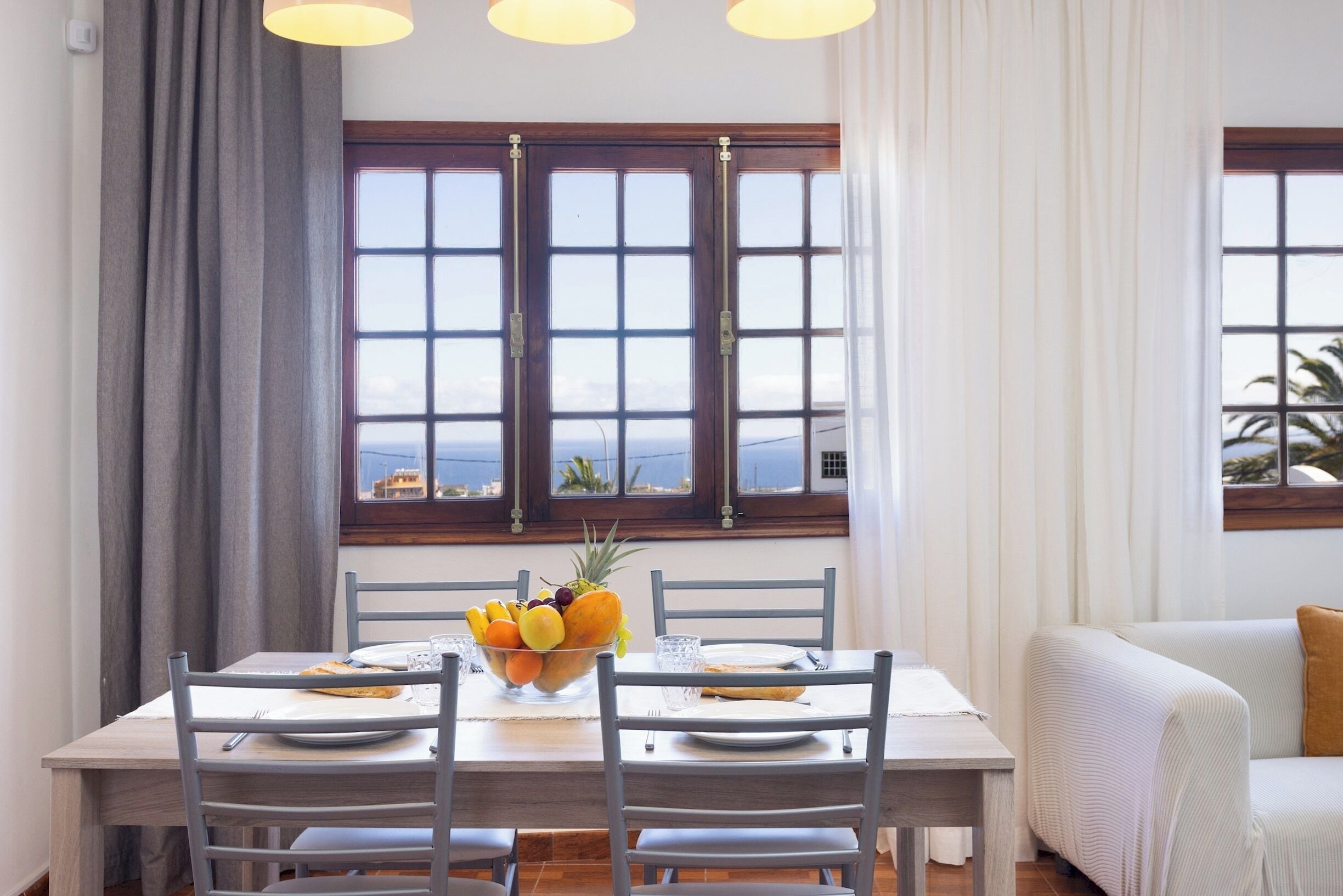 A dining area with a table set for two, a bowl of fruit, and a view of the sea through large windows.