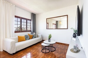 A modern living room with a sofa, a round coffee table, a TV mounted on the wall, and a window with curtains. - TenoHomes YSLA Escape Buenavista (Buenavista del Norte)