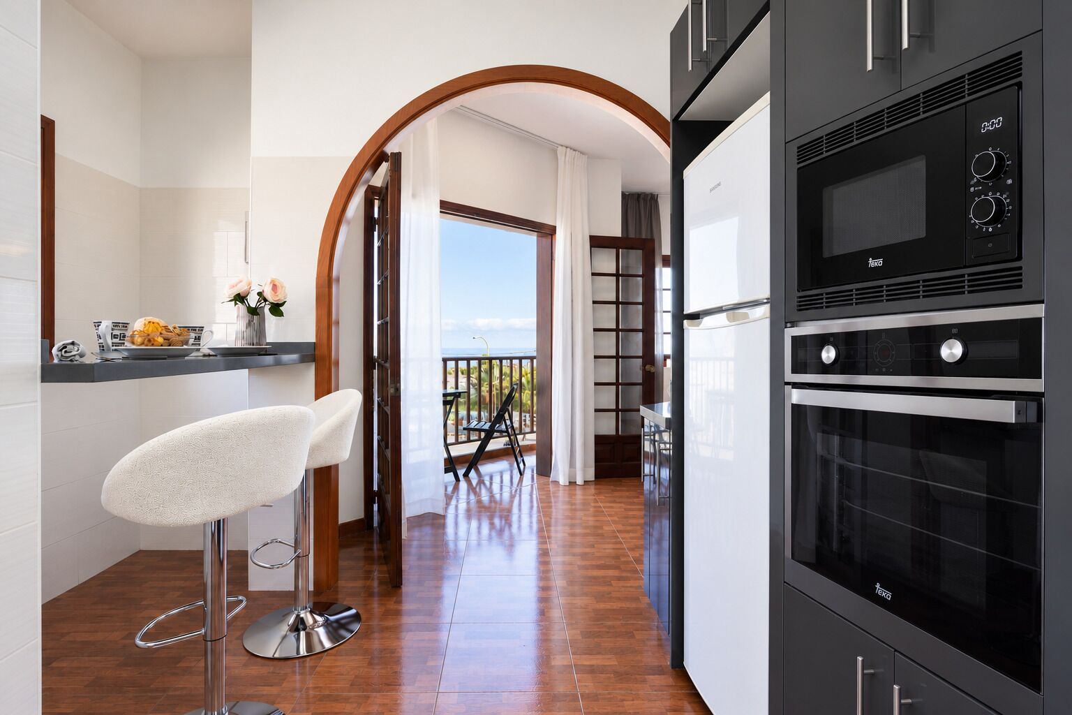 A modern kitchen with a built-in oven and microwave, a bar area with a stool, and a view to a balcony.