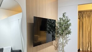 A modern living room with a flat-screen TV mounted on a wooden panel wall, a white console table with decorative items, a black sofa, and a large mirror. - Fully Furnished Makati Condo With Fast Wi-fi and Queen Bed (Makati)