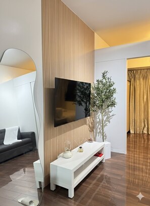 Ein modernes Wohnzimmer mit einem Flachbildfernseher an einer Holzpaneelwand, einem weißen Konsolentisch mit Dekorationen, einem schwarzen Sofa und einem großen Spiegel.