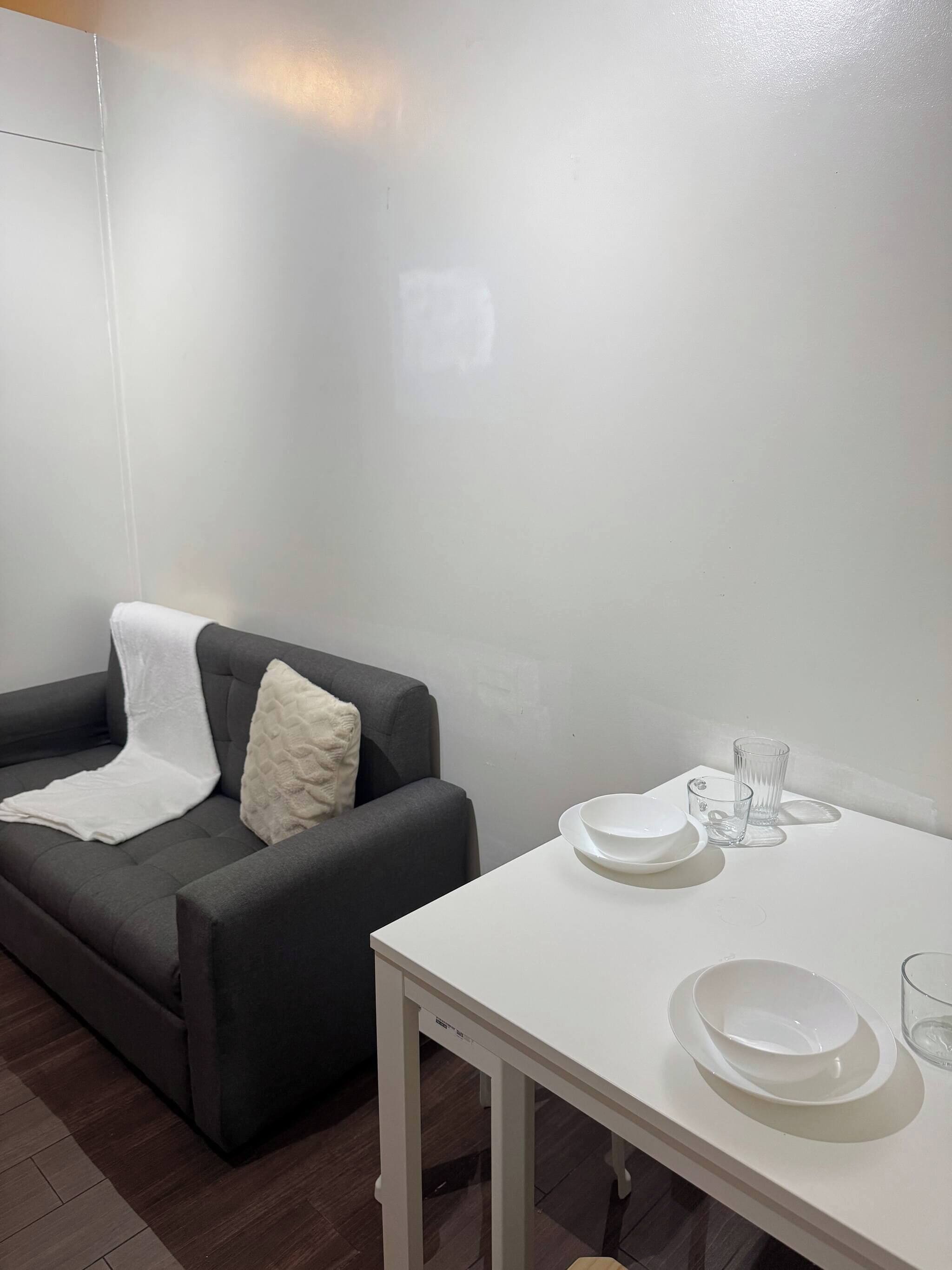 A room with a grey sofa, a white table with plates and glasses, and a white pillow.
