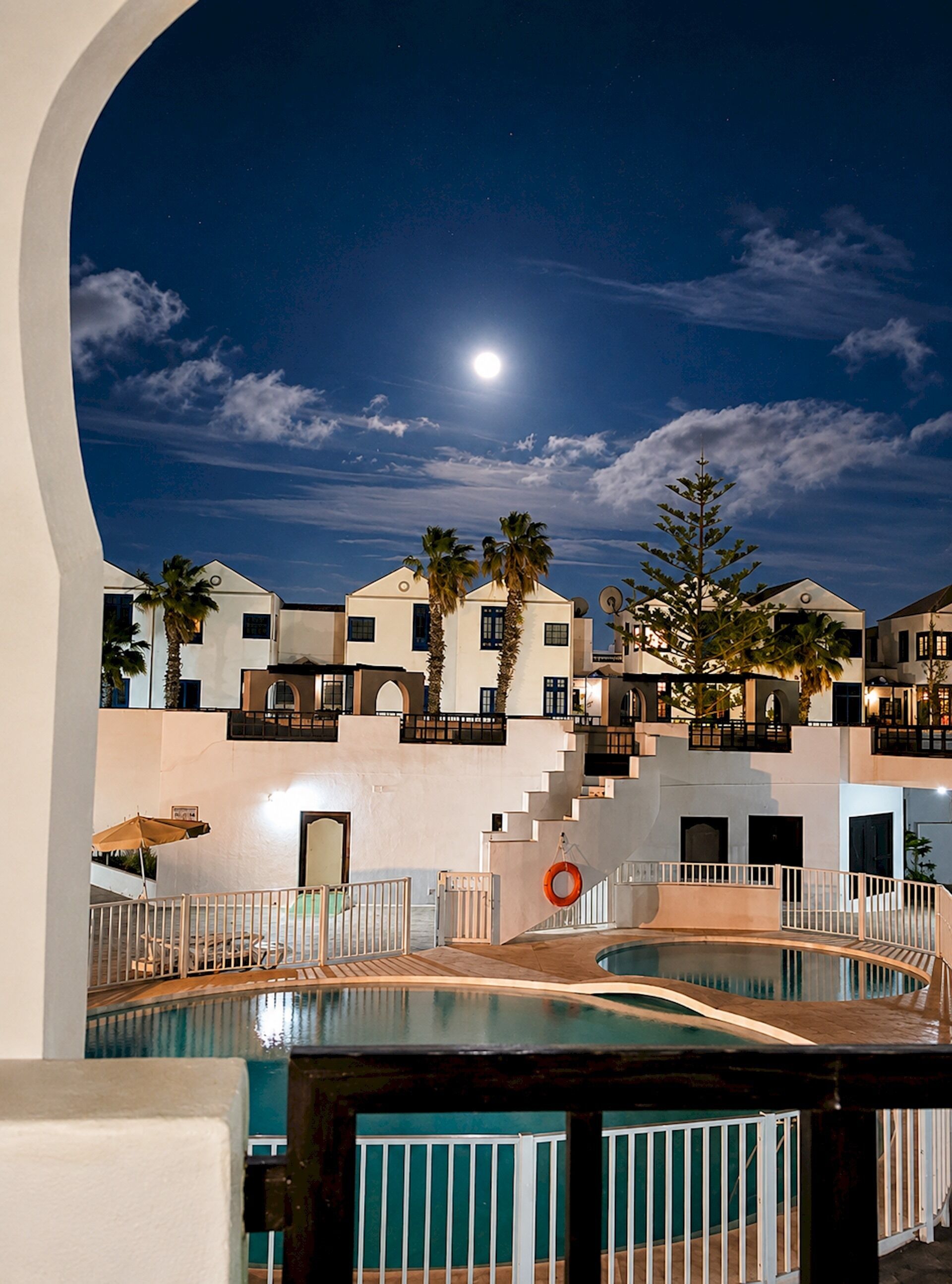 A nighttime view of a hotel with a swimming pool, palm trees, and a moon in the sky.