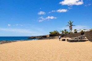 A beach with a clear sky, rocky shoreline, and a person walking. - Home2Book Golden Apt Pool & Terrace Costa Teguise (Costa Teguise)