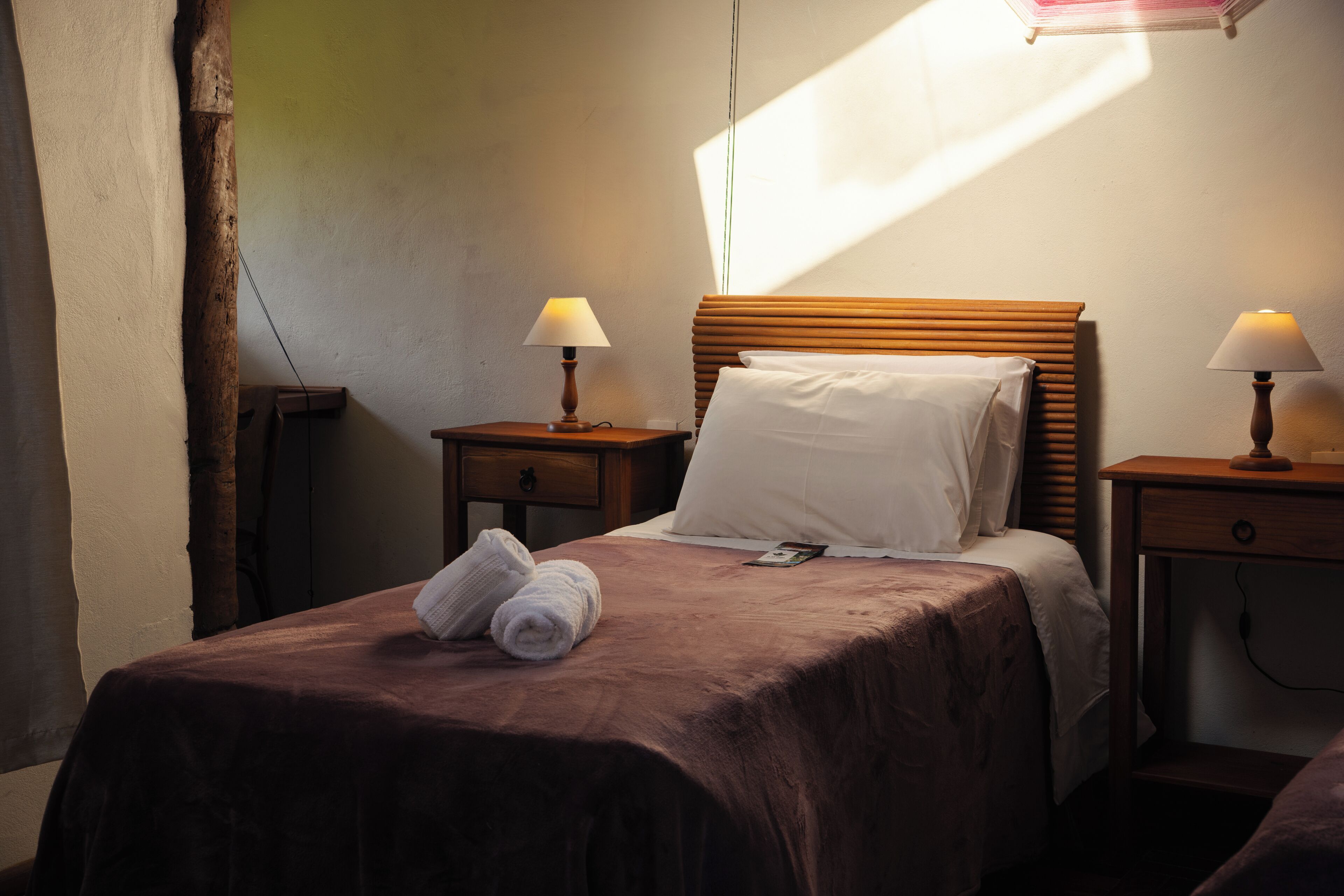 A hotel room with a bed, two bedside tables with lamps, a wooden headboard, a window with curtains, and a pair of towels on the bed.