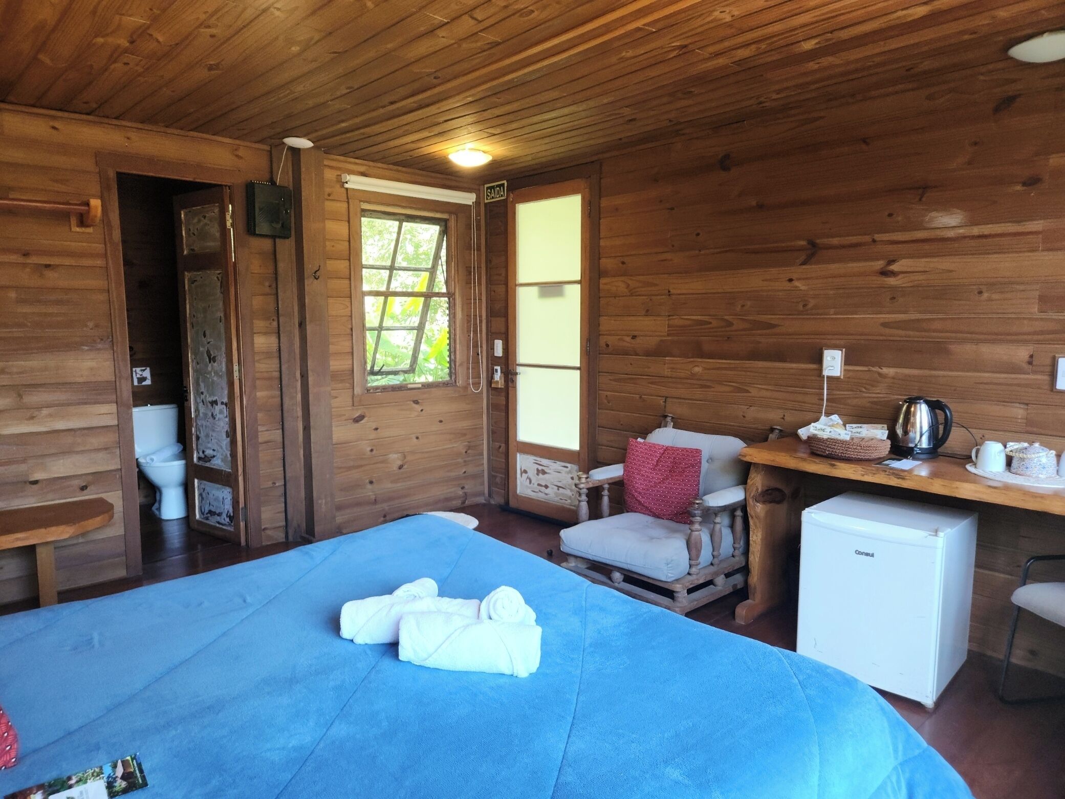 A wooden cabin room with a bed, a small table, a chair, a desk, and a refrigerator.