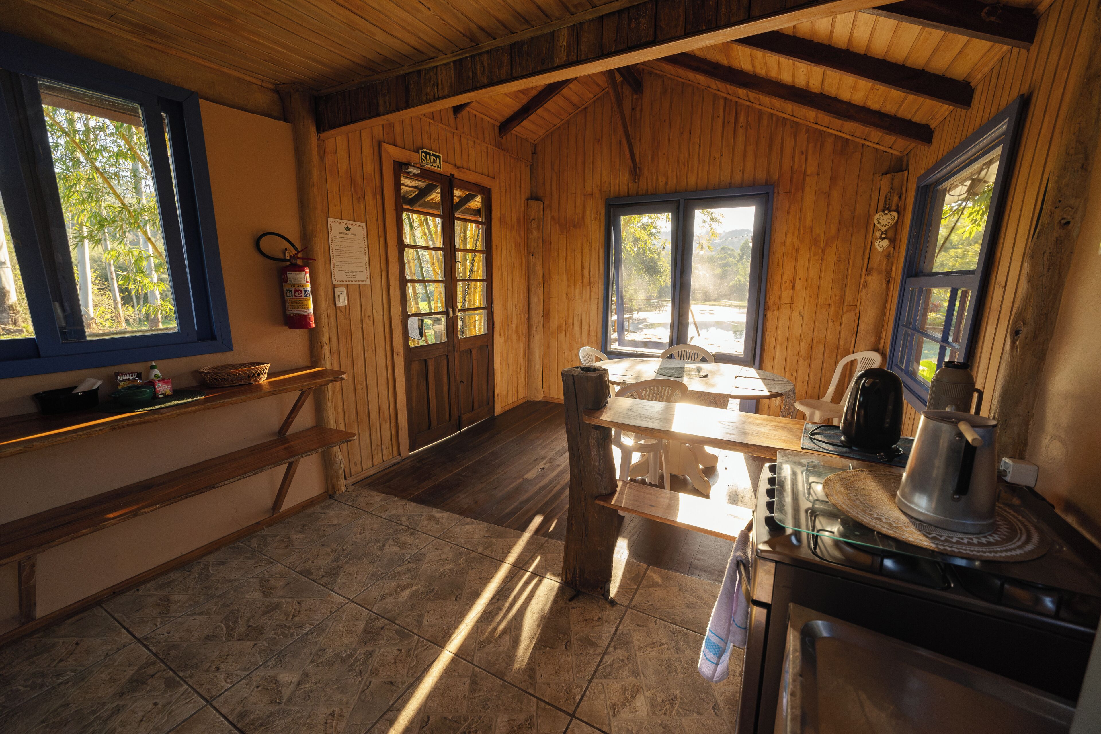 A rustic kitchen with wooden walls, a tiled floor, a stove, and a window overlooking greenery.