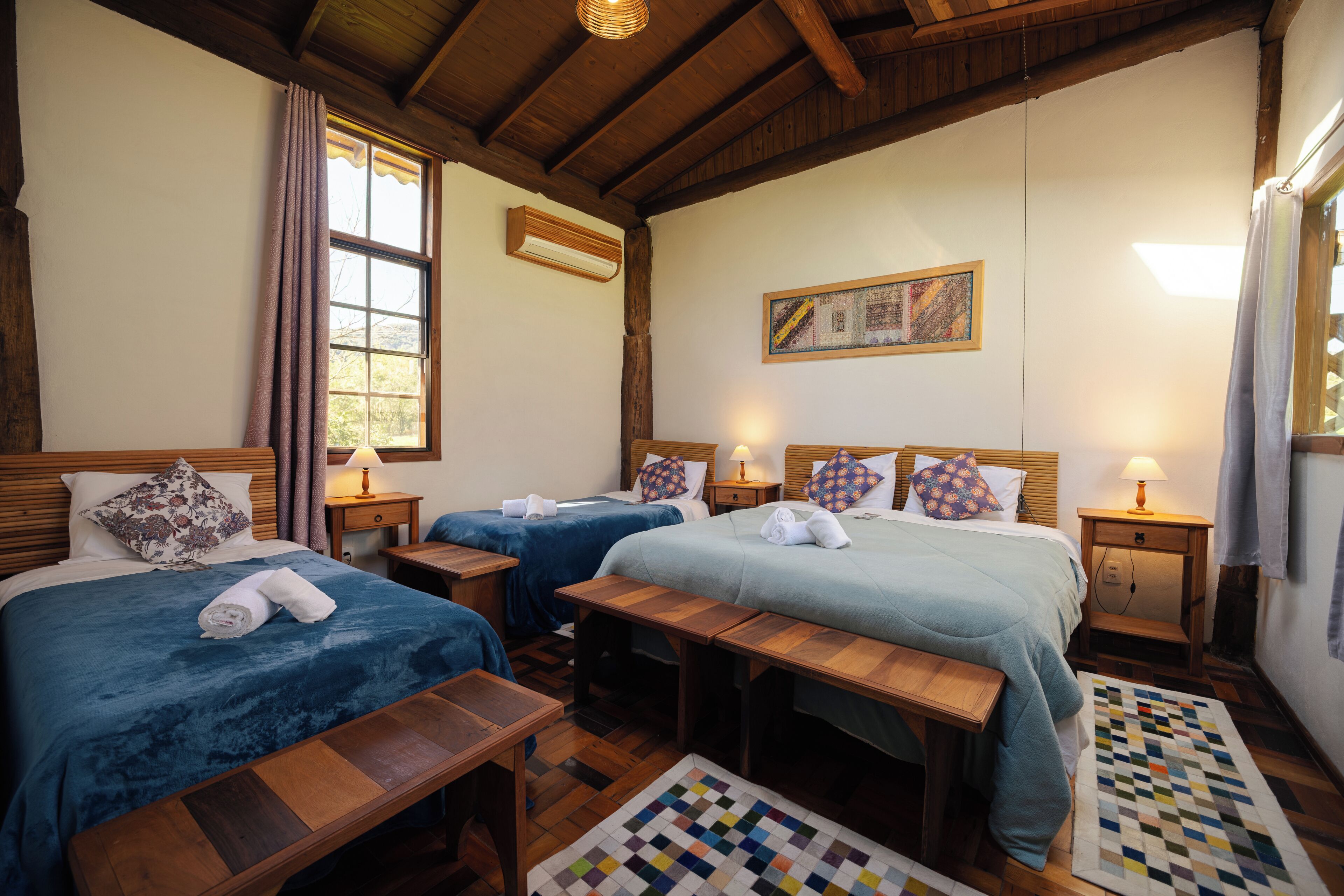 A hotel room with two beds, a wooden ceiling, a quilted rug, and a painting on the wall.