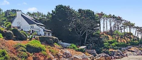 Uma propriedade costeira com uma casa grande, praia de pedras e árvores densas.