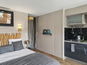 A compact hotel room with a bed, bedside lamp, wall-mounted light, a small shelf with books, a striped curtain, and a kitchenette with a microwave and utensils. - Cozy studio for 2 people — Les Villas de la Plage, Rospico (Névez) (Névez)