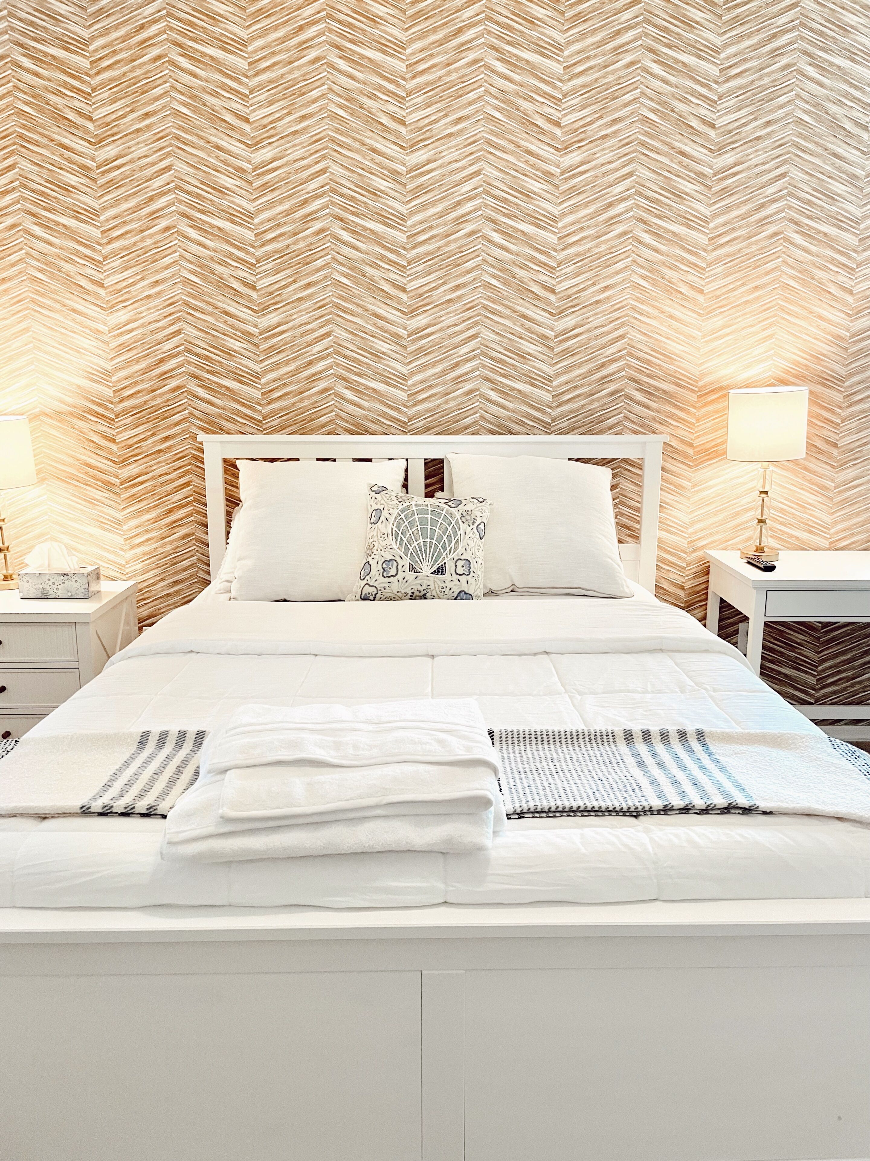 Un lit bien fait, avec du linge de lit blanc et un coussin à motifs, une table de chevet avec une lampe, et un mur orné d’un motif en chevrons.