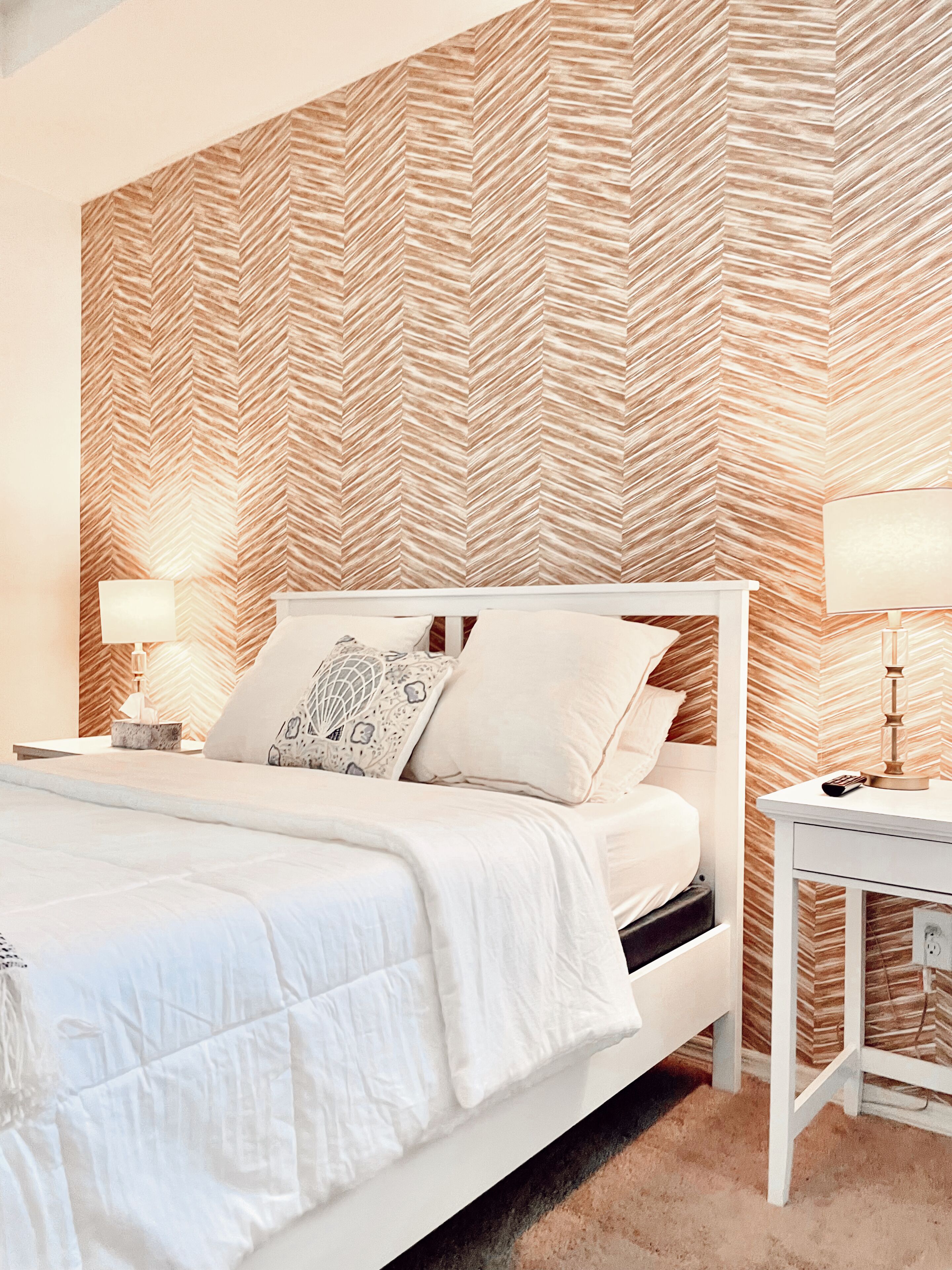 A bedroom with a herringbone-patterned wallpaper, a white bed with multiple pillows, and bedside tables with lamps.