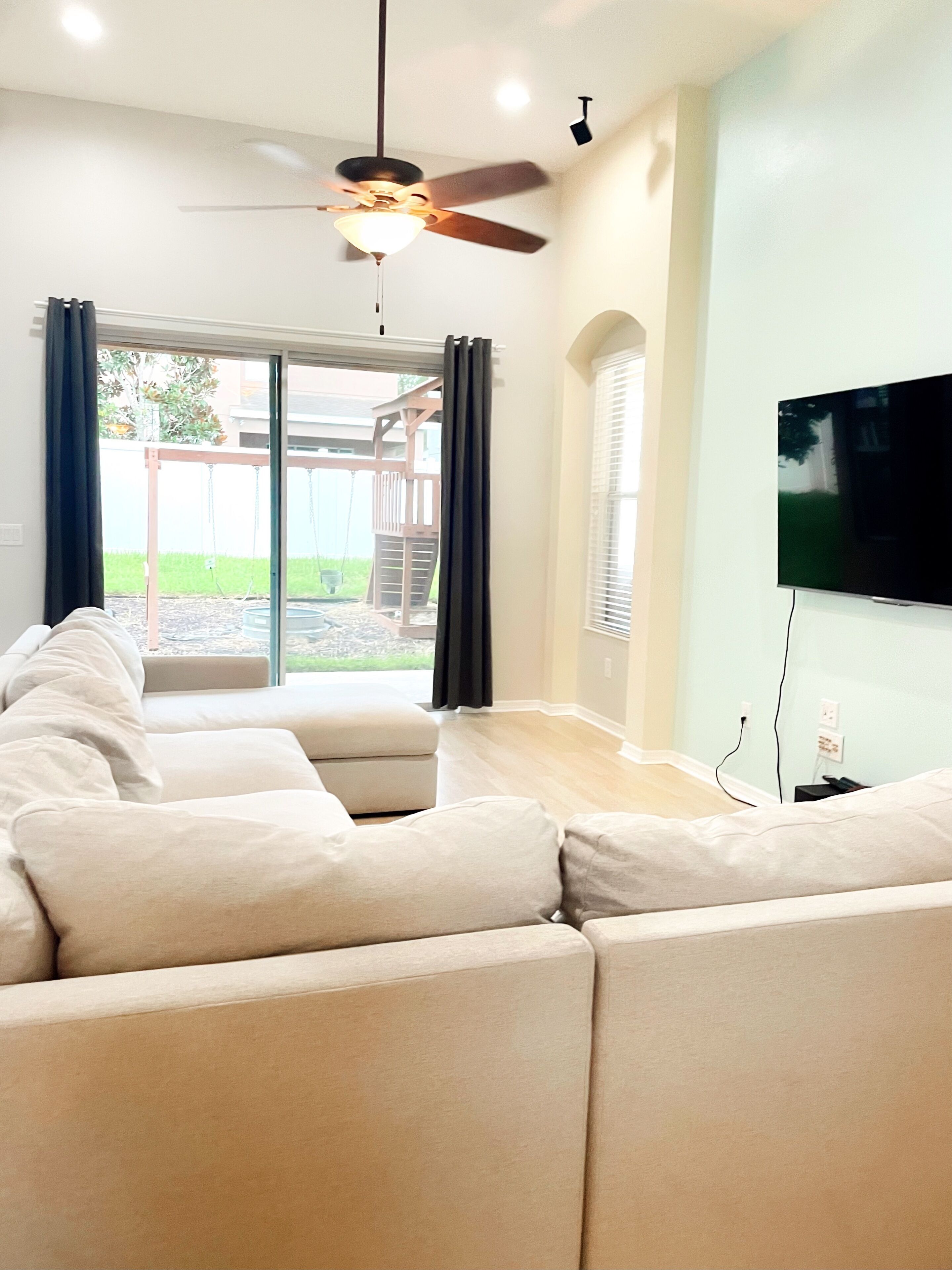 A living room with a sectional sofa, a ceiling fan, a television, and a view of the outdoors.