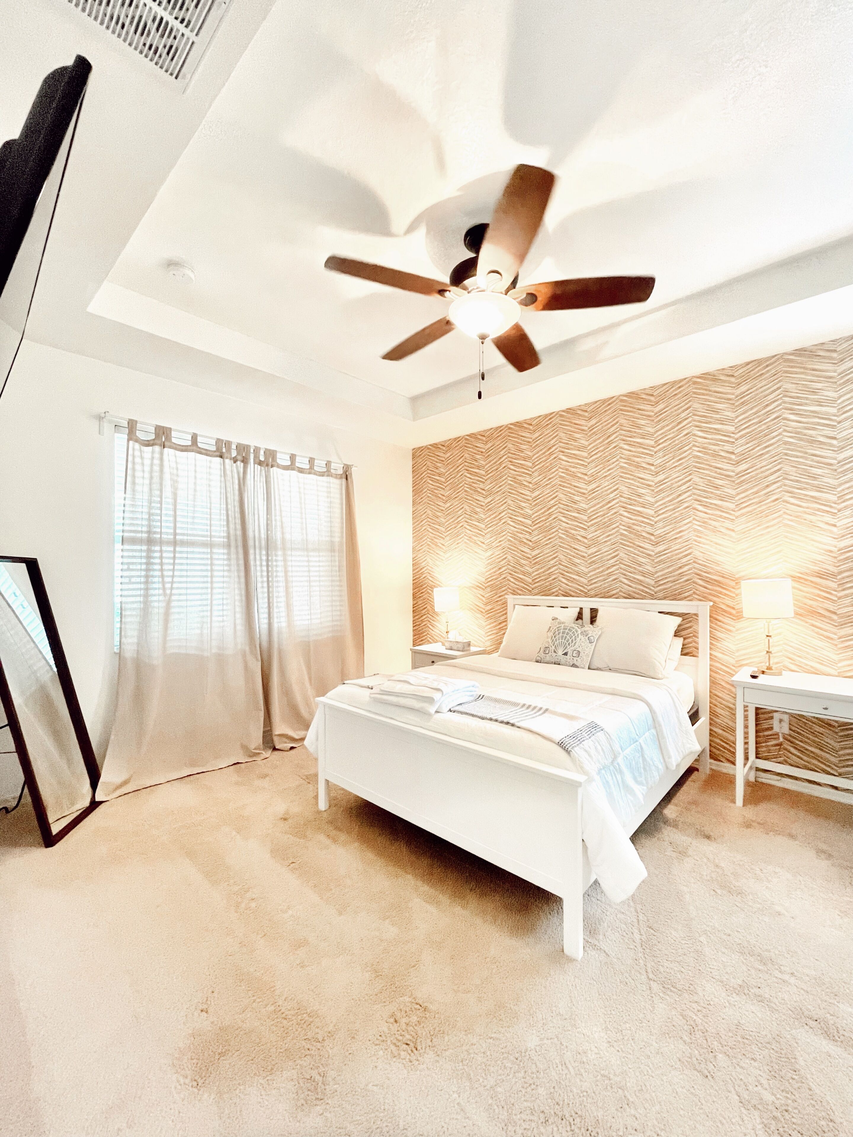 A bedroom with a bed, a ceiling fan, a bedside table with a lamp, and a wall with a herringbone pattern.