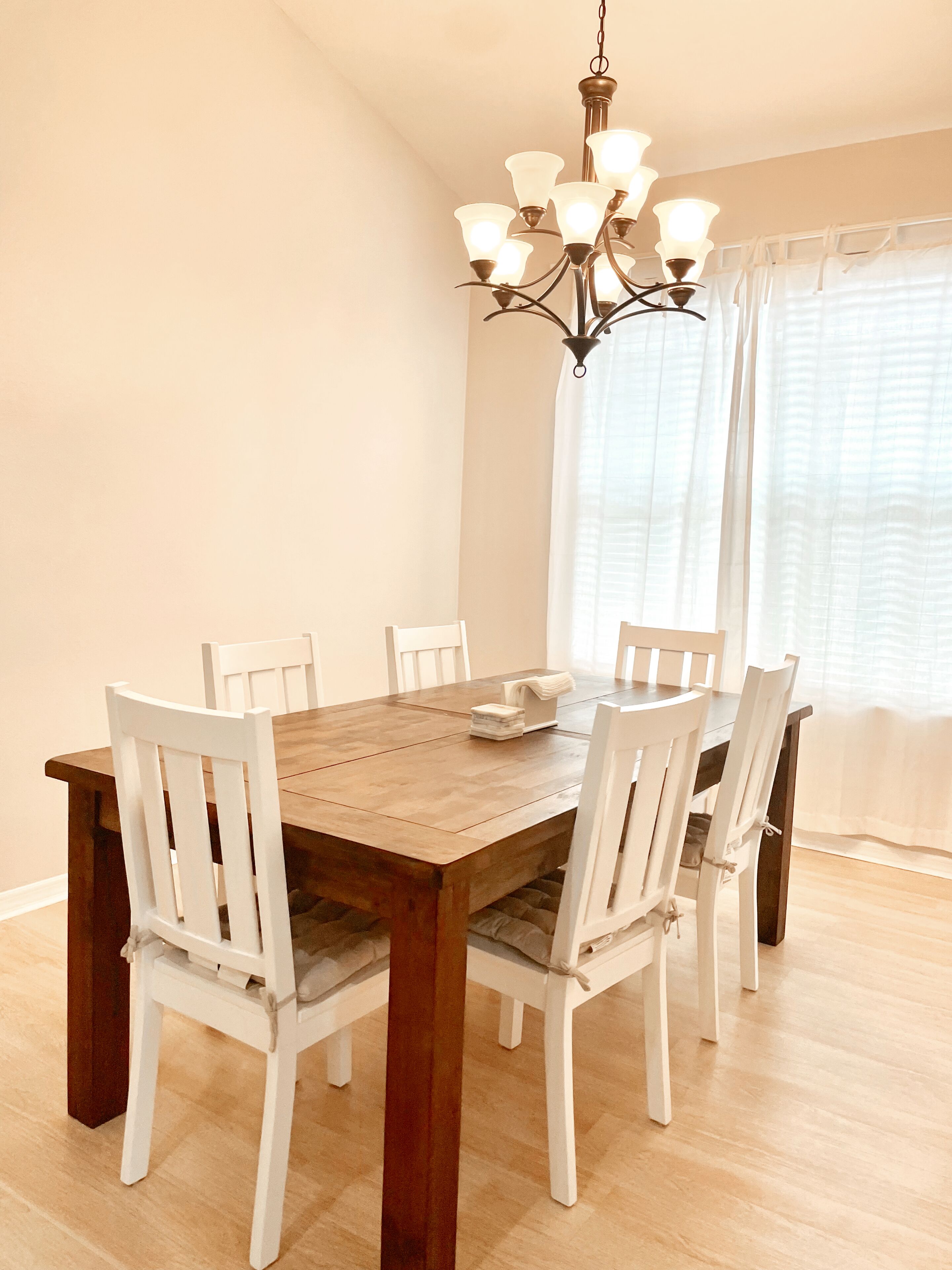A dining area with a wooden table and white chairs, a chandelier, and a window with curtains.