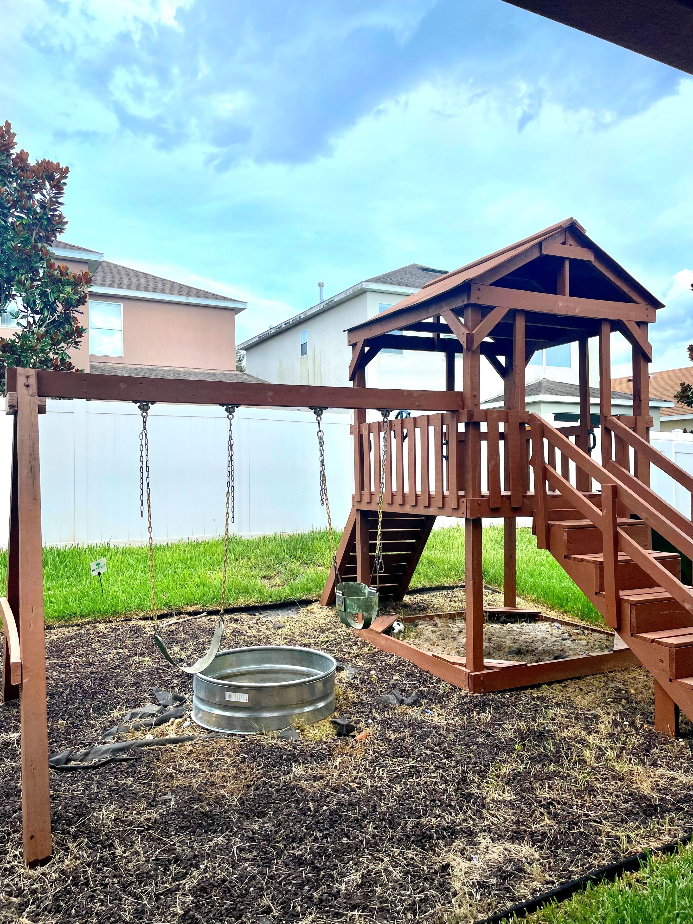 A wooden swing set with a metal bucket swing and a wooden playhouse with stairs.
