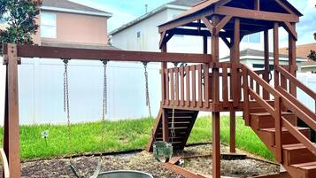 A wooden swing set with a metal bucket swing and a wooden playhouse with stairs.