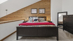 Une chambre avec un plafond en pente, un lit recouvert d’une couverture rouge, deux tableaux encadrés accrochés au mur et une commode.