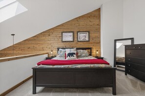 Ein Schlafzimmer mit schrägen Wänden, einem Bett mit roter Decke, zwei gerahmten Bildern an der Wand und einer Kommode.