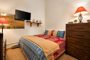 A bedroom with a bed, a dresser, a television mounted on the wall, and a lamp. - Cozy Loft Condo Near Granby Ranch | Pool, Hot Tubs & Mountain Access (Granby)
