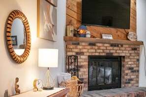 A living room with a brick fireplace, a mounted television, a wooden-framed mirror, and a table with a lamp and clock. - Cozy Fraser Retreat Near Winter Park Skiing (Fraser)