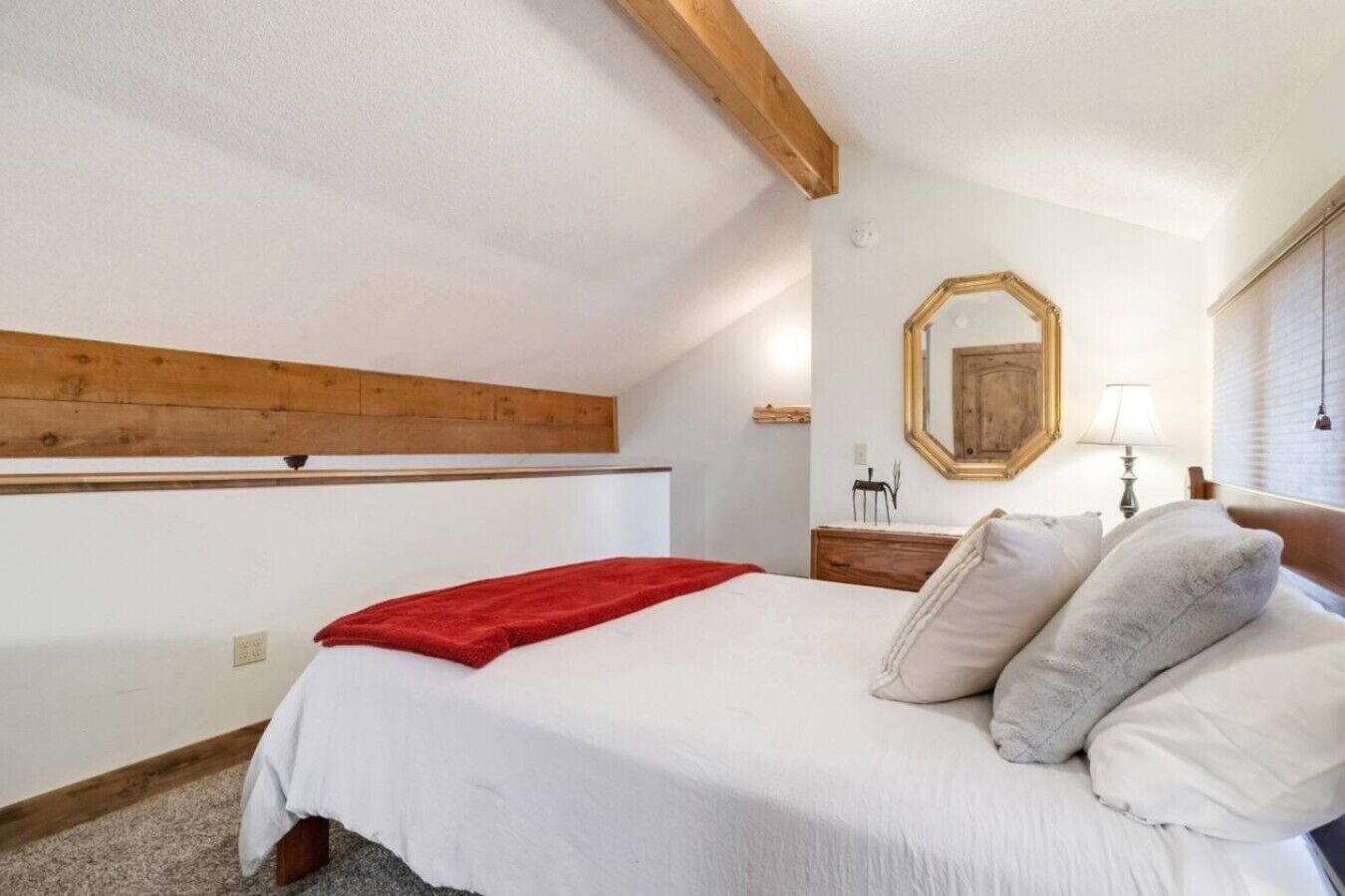 Een slaapkamer met een bed, een rode plaid, een houten plafondbalk, een houten dressoir, een spiegel en een raam met jaloezieën.