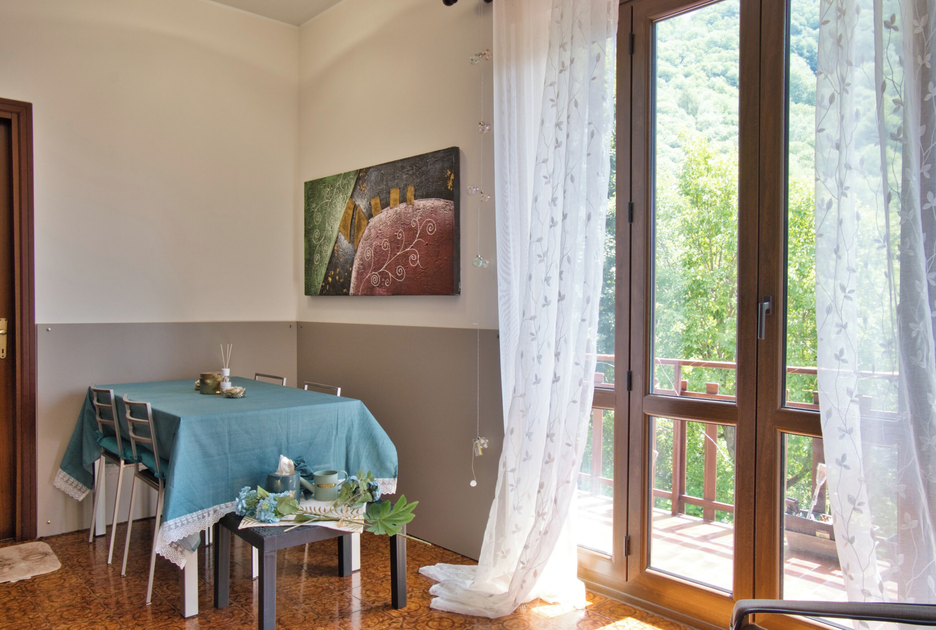 Ein Essbereich mit Tisch für zwei Personen, einem Bild an der Wand und Blick auf einen Balkon mit viel Grün.