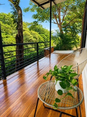 Um deck de madeira com guarda-corpo de metal, uma cadeira de vime e um vaso com planta.