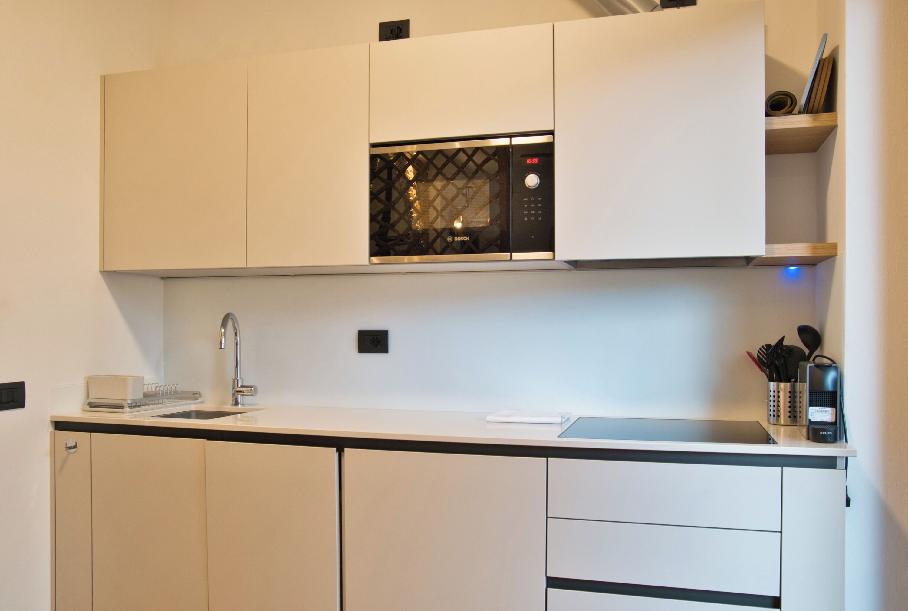 Une cuisine moderne avec des meubles blancs, un four à micro-ondes de la marque Bosch, un évier et un plan de travail sur lequel se trouvent une machine à café et des ustensiles.